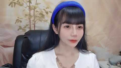 Media: A video of an East Asian woman with long black hair, fair skin, and a blue headband, wearing a white top and sitting in a black chair against a beige, leafy wallpaper backdrop.