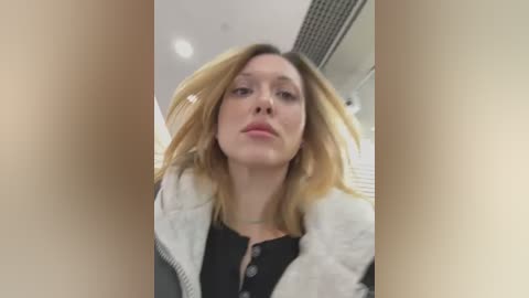 Media: A video of a blonde woman with light skin, wearing a black top and white fur-trimmed jacket, taken from a low angle in an indoor setting with white walls and a grid ceiling.