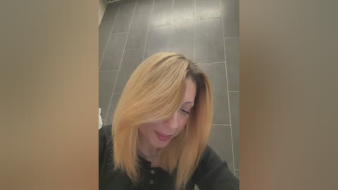 Media: Video of a smiling, fair-skinned woman with straight, shoulder-length blonde hair, wearing a black shirt. She is standing in a modern, tiled hallway with a beige wall and grey floor tiles.