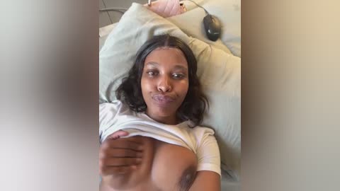 Media: Video of a Black woman with dark skin and long, wavy black hair, lying on a bed, lifting her white t-shirt to reveal her medium-sized breasts.