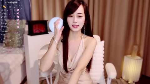 Media: A video of a slender Asian woman with long dark hair, wearing a beige halter dress, seated on a white chair. She holds a white orb. Background features a TV screen, curtains, and a white table.