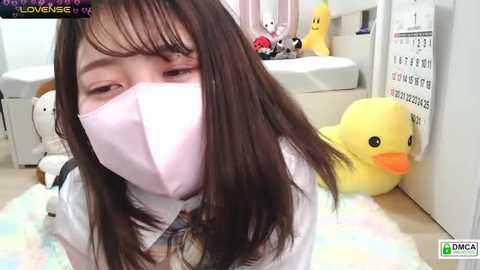 Media: Video of an Asian woman with long brown hair, wearing a white mask and white shirt, seated in a colorful, playful room with stuffed toys and a calendar.