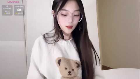 Media: Video of an East Asian woman with long black hair, wearing a cozy white sweater with a teddy bear design, and clear glasses, sitting in a minimalist, light-colored room.