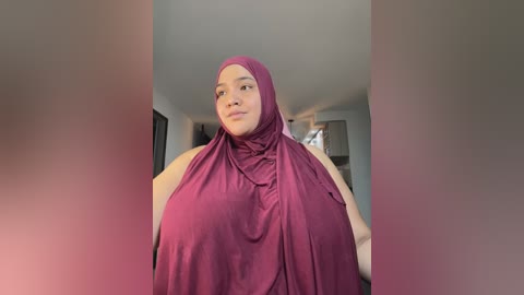 Media: Video of a plus-size woman in a sleeveless, maroon hijab, standing in a minimalist, well-lit room with muted, neutral-colored walls.