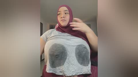 Media: A video of a plus-sized woman in a maroon hijab and gray shirt with large gray boob prints, posing in a hallway with blurred background.