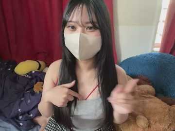 Media: A video of an Asian woman with long black hair, wearing a beige mask, a gray tank top, and checkered skirt, surrounded by stuffed toys in a messy bedroom with red curtains.