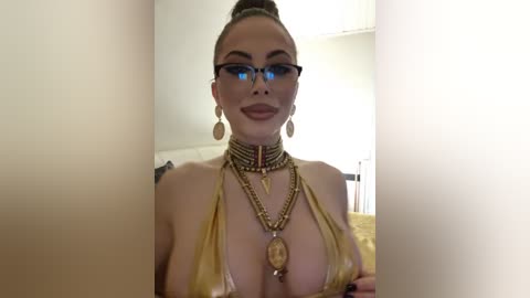 Media: Video of a fair-skinned woman with a high bun, wearing blue-framed glasses, a gold bikini, gold jewelry, and a gold choker with a pendant. She has large earrings and is smiling.