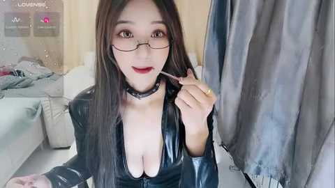 Media: A video of an Asian woman with long black hair and glasses, wearing a black latex dress, holding a lollipop, in a modern living room.