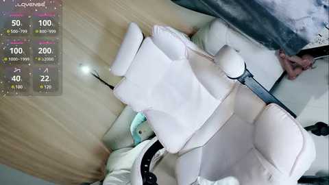 Media: Video of a modern, light-colored office chair with white cushions, viewed from above. The background shows a wooden floor and a person, partially visible, wearing a white shirt. A digital display in the top left corner shows temperature and humidity readings.