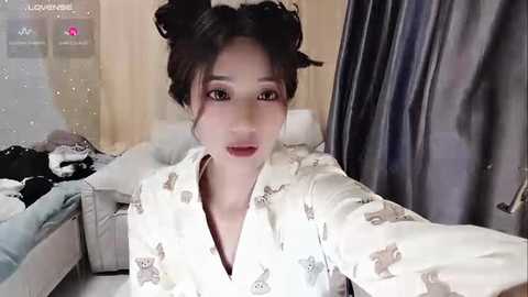 Media: A video of a young East Asian woman with fair skin, wearing a white kimono adorned with deer patterns, taking a selfie. She has dark hair styled in two buns. Background shows a cluttered bedroom with wooden furniture and a bed.