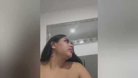 Media: A video of a nude Latina woman with long dark hair, medium skin tone, and full lips, looking upward, in a bathroom with a mirror and white walls.