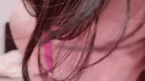 Media: A close-up video of a woman's shoulder and upper arm, with her dark brown hair partially covering her face and a vivid pink earring visible. The background is blurred, focusing on the hair and earring.