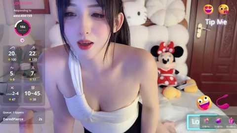 Media: Video of an East Asian woman with light skin, wearing a white strapless top, posing in a room with plush toys and a Minnie Mouse doll.