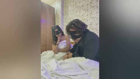 Media: Video of a woman with long black hair and a man with shoulder-length brown hair, both wearing black masks, sitting on a bed with white sheets, in a dimly lit room with patterned wallpaper.