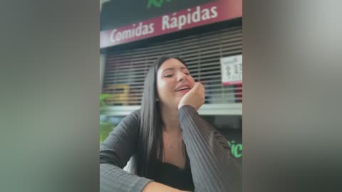 Media: Video of a Latina woman with long black hair, wearing a grey sweater, sitting in a convenience store with a snack display behind her.