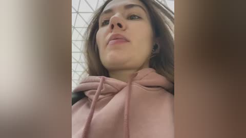 Media: Video of a young woman with fair skin and brown hair, wearing a pink hoodie, gazing upwards with a neutral expression. Background features a geometric patterned ceiling.