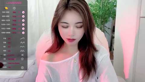 Media: A young Asian woman with long, straight brown hair, wearing a white off-shoulder blouse, sits in a room bathed in soft pink light.