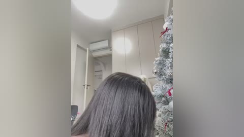 Video of a woman with long, straight dark hair, facing away, in a modern, minimalistic room with white walls, a Christmas tree, and a white cabinet.