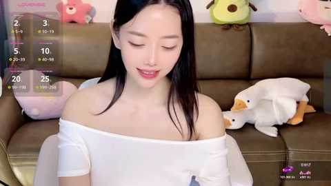 Media: A video of an East Asian woman with long black hair, wearing an off-shoulder white top, sitting on a brown leather couch with stuffed animals.