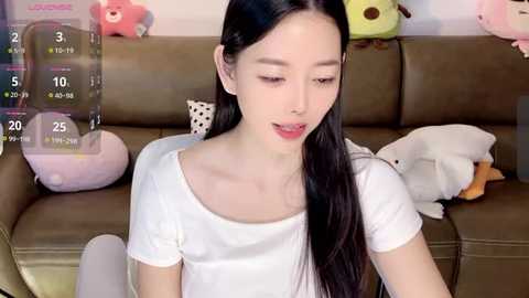 Media: A video of a young Asian woman with long black hair and fair skin, wearing a white t-shirt, sitting on a brown couch with stuffed animals.