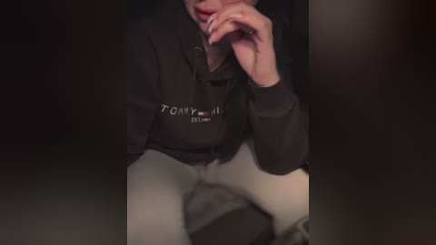 Media: A dimly lit video of a woman in a black hoodie and white pants, with her fingers near her lips, creating a mysterious and intimate atmosphere.