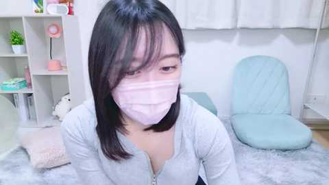 Media: Video of a young Asian woman with shoulder-length black hair, wearing a light gray zip-up sweater and a pink face mask, sitting on a light blue chair in a minimalist room with white walls and a fluffy white rug.