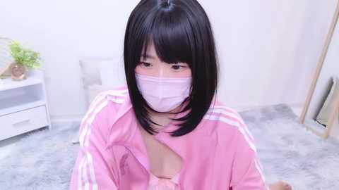 Media: Video of an East Asian woman with short black hair, wearing a pink tracksuit jacket with a revealing neckline, white face mask, and a gray carpeted room.