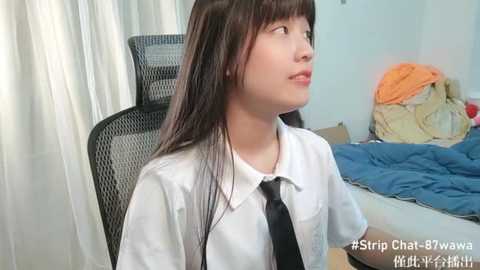 Media: A video of a young Asian woman with long black hair, wearing a white shirt and black tie, sitting in a black mesh chair in a messy bedroom with white curtains and a bed with blue sheets.