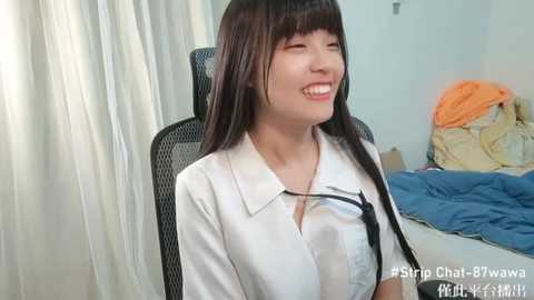 Media: A video of a smiling Asian woman with long, straight black hair and pale skin, wearing a white blouse, sitting in a black mesh chair. The background shows an unmade bed with blue and orange blankets, and white curtains.