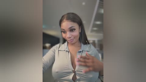 Video of a young Black woman with medium brown skin, straight black hair, and large breasts, wearing a low-cut gray blouse, smiling at the camera, taken in a modern indoor setting.