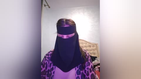 Media: Video of a person wearing a black niqab with a pink band, leopard print shirt, in a dimly lit room with a beige headboard and white walls.