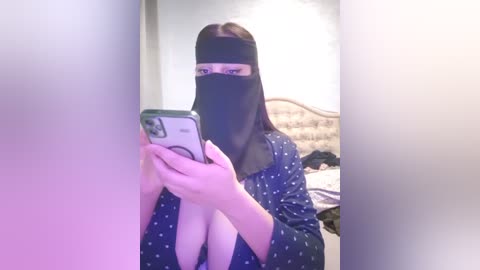 Media: Video of a woman with light skin, dark hair, and a black face veil, holding a phone, standing in a dimly lit bedroom with a bed and a partially visible headboard.