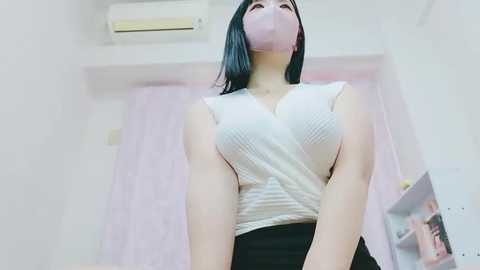 Media: Video of an Asian woman with fair skin and shoulder-length black hair, wearing a white, low-cut top and a black skirt. She has a pink face mask and is standing in a pastel-pink room with an air conditioner and a white shelf in the background.