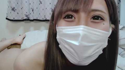 Media: Video of an Asian woman with long, straight brown hair and a surgical mask covering her mouth, lying on a bed with white sheets, in a room with a patterned curtain.