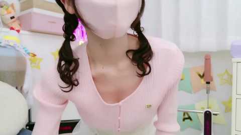 Media: Video of an Asian woman with light skin, wearing a pink mask, pastel pink ribbed sweater, and white pants, in a pastel-colored room with a bed, toys, and a pink vibrator.