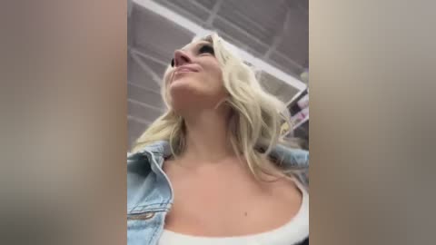 Media: Video of a blonde woman in a white tank top and denim jacket, smiling upward, against a white ceiling with exposed ductwork, creating a casual, candid indoor setting.