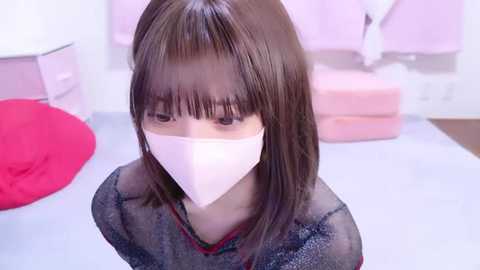 Media: A video of a young Asian woman with straight, shoulder-length brown hair, wearing a light pink mask, a sheer black top, and a red bra. She stands in a pink-tiled bathroom with a red stool and pink towels.