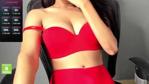 Media: Video of a woman with light skin, long black hair, wearing a vibrant red strapless bra and matching high-waisted shorts, sitting on a black mesh office chair. Background includes a gray wall and a TV screen displaying a \"LOVENSE\" logo.