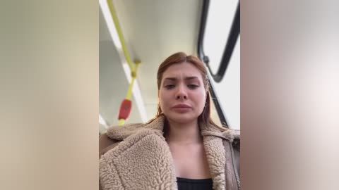 Media: Video of a young Caucasian woman with fair skin and straight, shoulder-length light brown hair, wearing a beige fleece jacket, seated on a bus with a yellow pole and red emergency stop sign in the background.