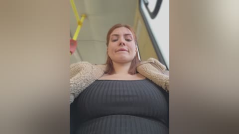 Media: Video of a Caucasian woman with shoulder-length red hair, wearing a black ribbed dress and beige cardigan, standing in a subway car.