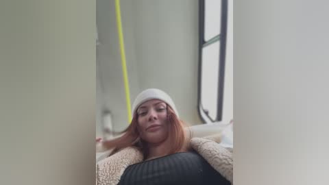 Media: Video of a young woman with fair skin, wearing a white beanie and black sweater, lying on a beige blanket in a dimly lit room with a large window and yellow lighting.