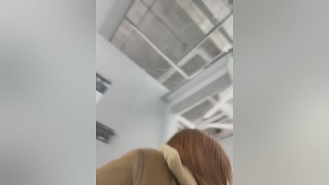 Media: A video capturing a blurred, side view of a person with long, auburn hair, wearing a beige jacket, walking in an industrial-style hallway with white walls and exposed concrete ceiling.