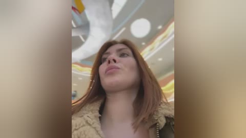 Media: Video of a young woman with fair skin and shoulder-length auburn hair, wearing a beige fur coat, looking up with a neutral expression in a brightly lit indoor setting with circular lights and colorful ceiling panels.