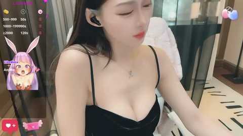 Media: Video of an Asian woman with long black hair and pale skin, wearing a black camisole revealing cleavage, sitting in a modern room.