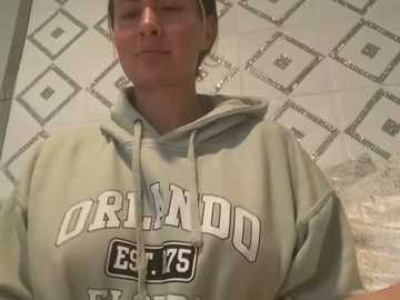 Media: Video of a woman with light skin and short brown hair, wearing a gray hoodie with \"ORDANDO EST 1975\" on the front, standing in front of a tiled wall with a geometric pattern.