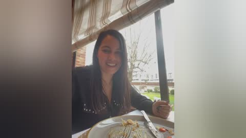 Media: Video of a smiling, light-skinned woman with long, straight dark hair, wearing a black shirt, sitting at a table with a half-eaten pizza. Background shows a window with striped shades, outdoor greenery, and a cloudy sky.