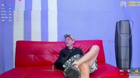 Media: Video of a young, smiling white man in a black t-shirt, camo shorts, and cap, reclining on a red leather couch against a purple wall. A black standing fan and VR headset are visible.