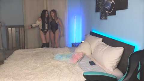 Media: Video of two women in black lingerie standing by a bed with white sheets, one holding a toy, in a modern, dimly lit bedroom with blue lighting and abstract art on the wall.