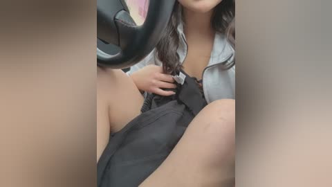 Media: Video of a woman sitting in a car, wearing a gray jacket and black skirt, with her seatbelt on, her legs slightly apart, showing her thigh.