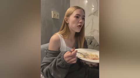 Media: A video of a young blonde woman in a white tank top and gray hoodie eating spaghetti with a fork, sitting in a dimly lit room with a beige wall and a white chair.